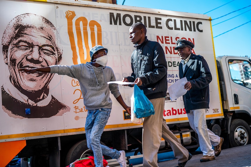 Someone directs two men towards something out of the frame. In the background is a truck sign which says "mobile clinic".