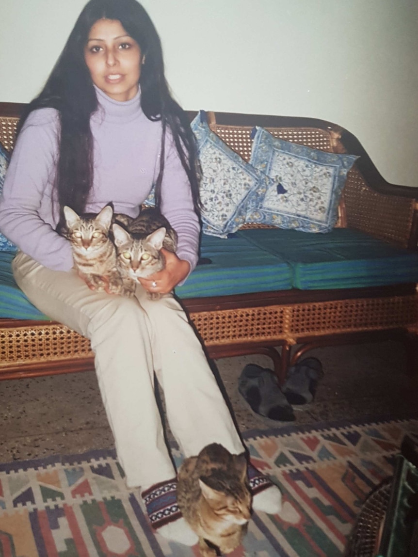 A young girl with long black hair sits on a rattan sofa with two cats on her lap and one at her feet.