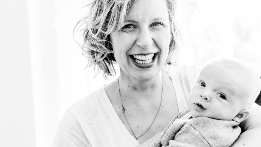 Black and white photo of Alexandra Collier smiling very widely holding a small baby in her arms, who looks at the camera.
