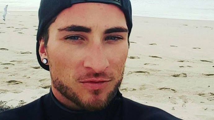 Matthew Fisher-Turner takes a seflie while standing on a beach wearing a black cap and jumper.