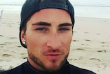 Matthew Fisher-Turner takes a seflie while standing on a beach wearing a black cap and jumper.