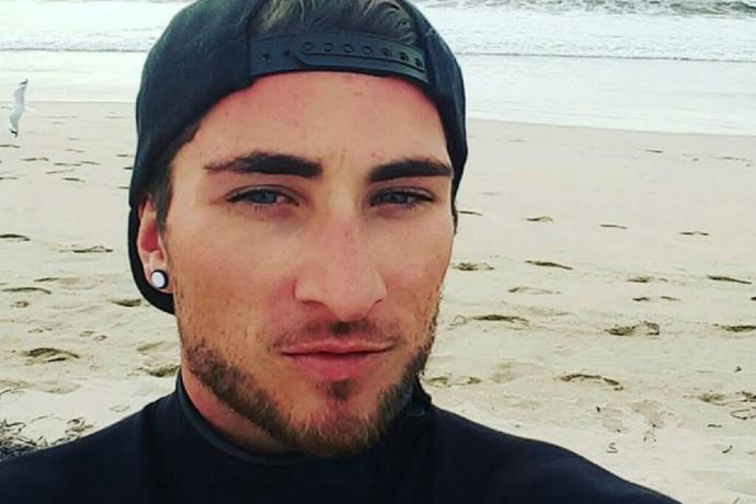 Matthew Fisher-Turner takes a seflie while standing on a beach wearing a black cap and jumper.