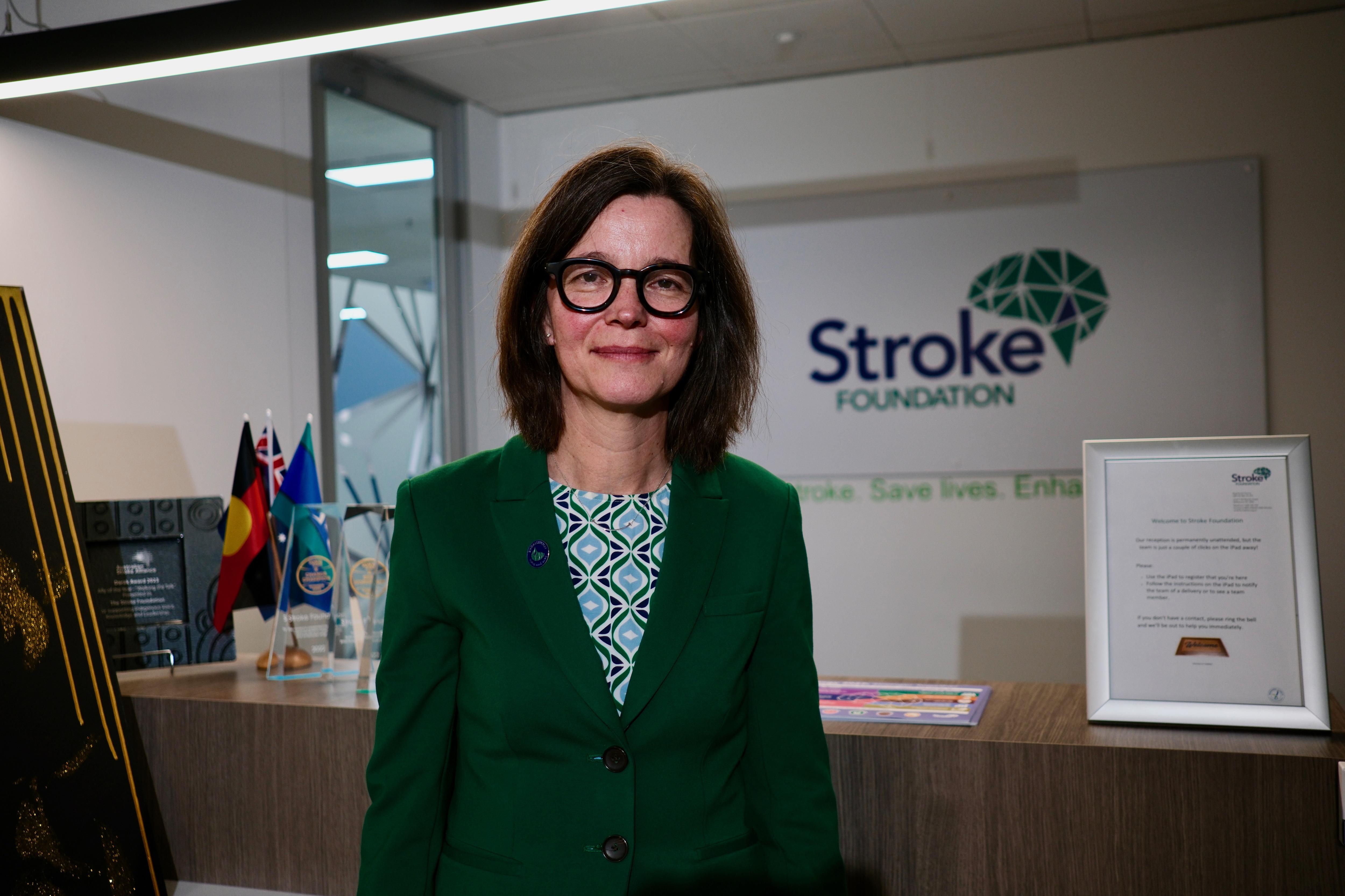 A woman with brunette hair wearing glasses, a dark green blazer and floral top stands in front of "Stroke Foundation" signage