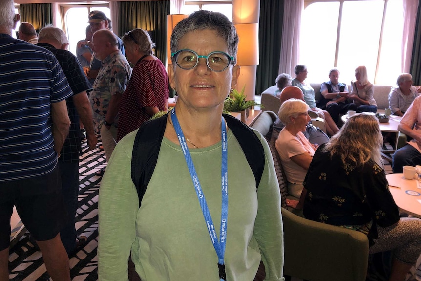 A woman stands in a lounge area on a cruise ship smiling for a photo wearing a green jumper and spectacles in front of others.