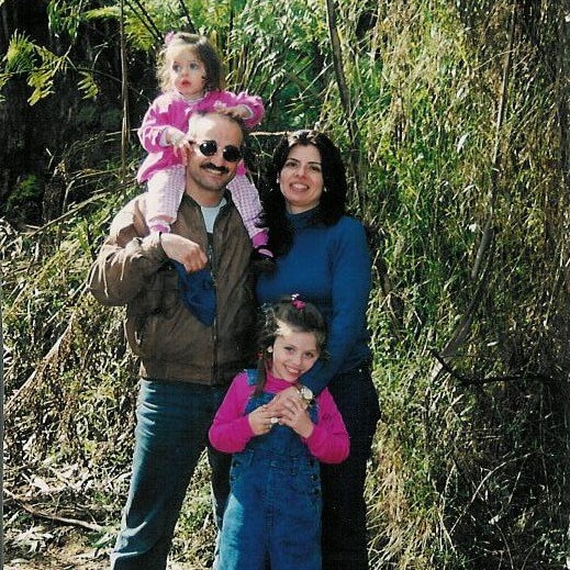 A man and woman stand smiling, a young girl stands in front of them and a toddler sits on the father's shoulders.