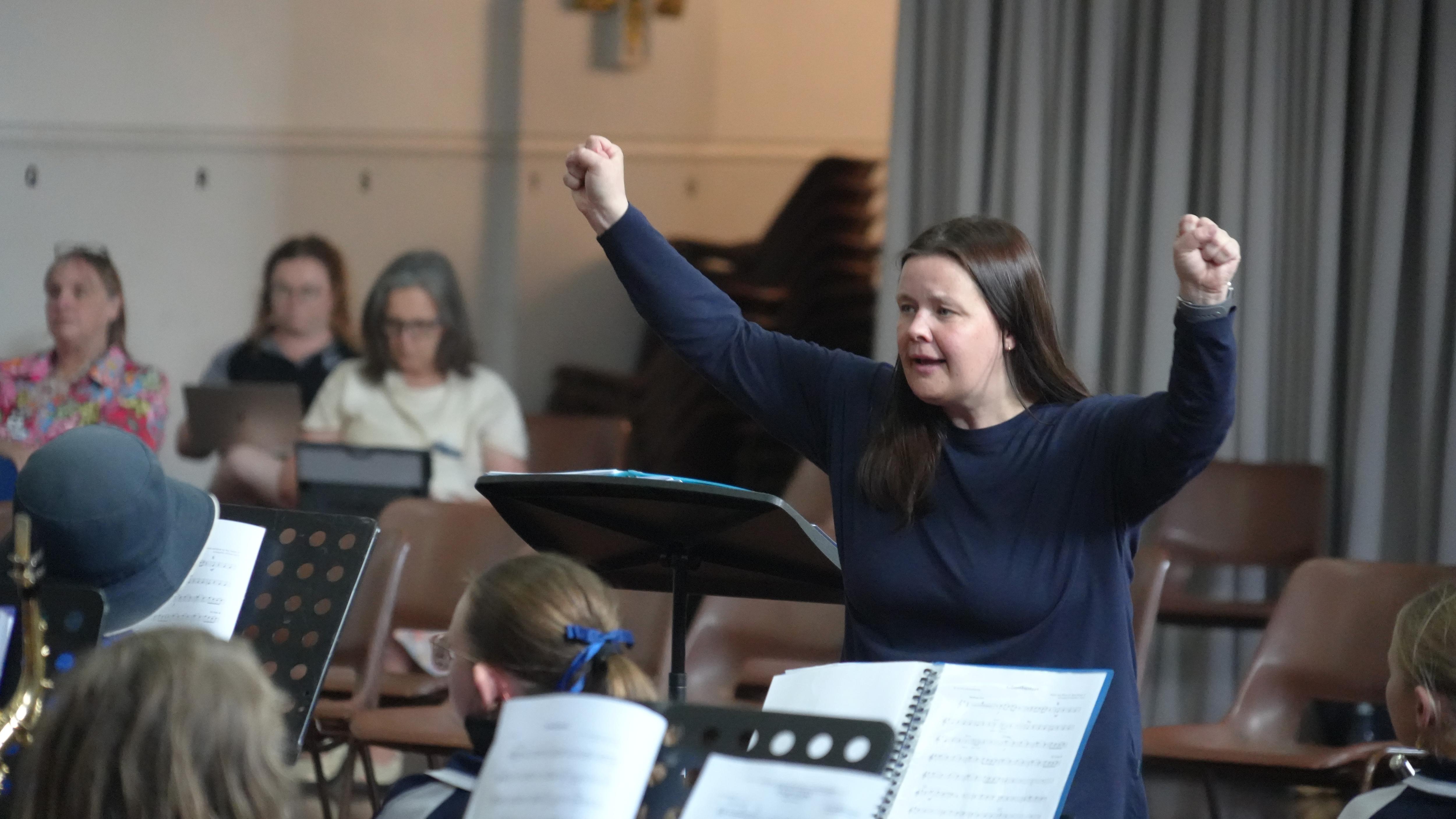 A brunette woman standing in front of a group of children playing instruments, her arms up to the sky.