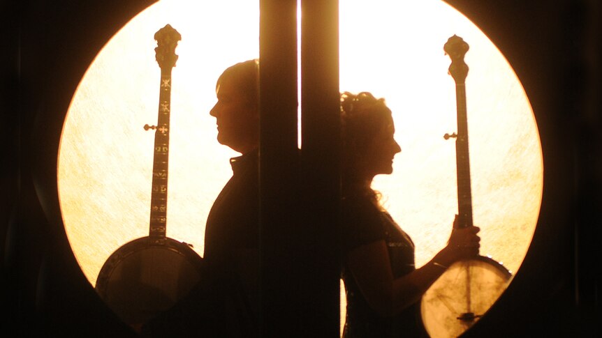 A man and a woman, both holding banjos, stand back to back with a circle of light behind them