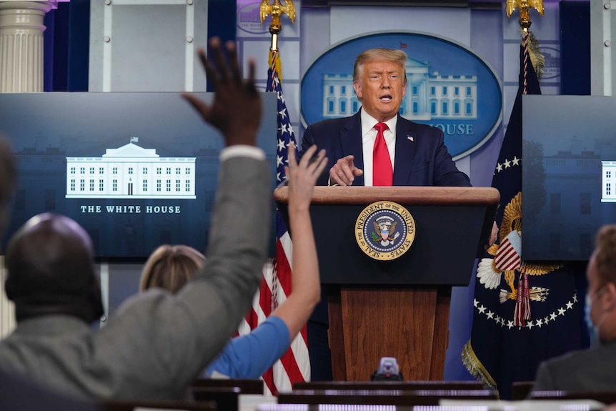 President Donald Trump speaks from behind a podium during a news conference, while reporters hold their hands up