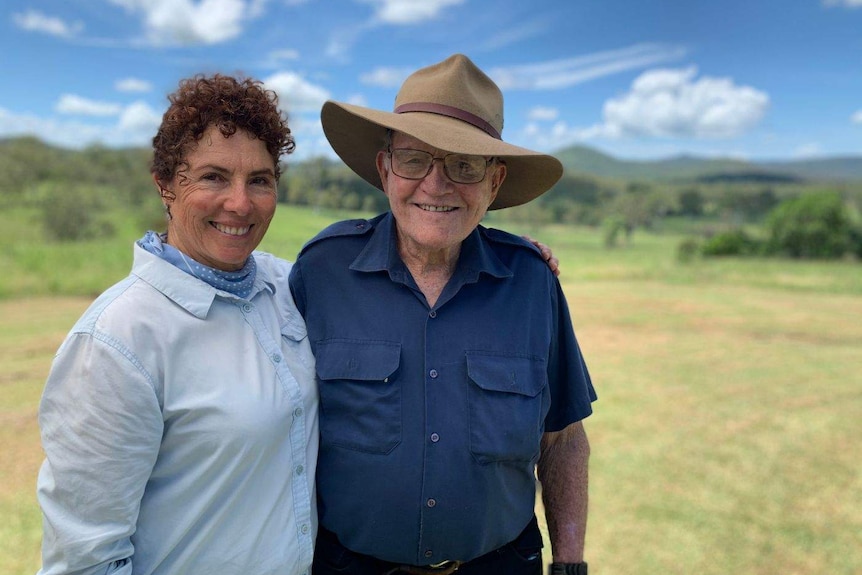 A middle-aged woman with red curly hair hugs an elderly man wearing a blue shirt and hat.