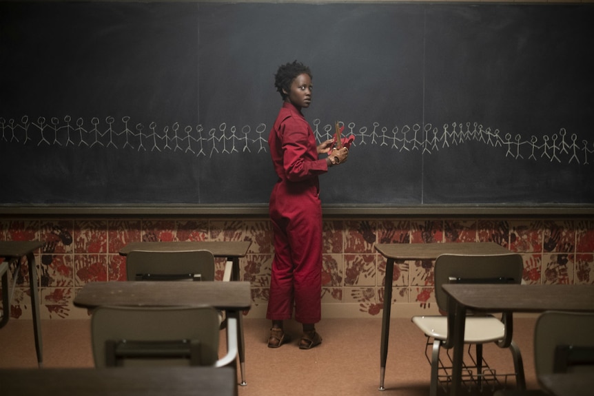 The character, wearing a red jumpsuit, stands in front of a classroom black board which is covered in a drawing of stick figures