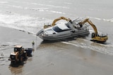 Two diggers taking apart a yacht in shallow waters at a beach, with a loader in front