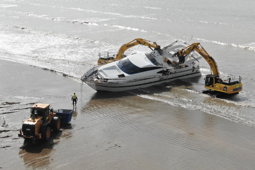 Two diggers taking apart a yacht in shallow waters at a beach, with a loader in front