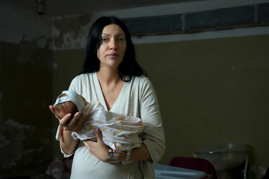 A woman in white dress with dark hair holds a newborn baby