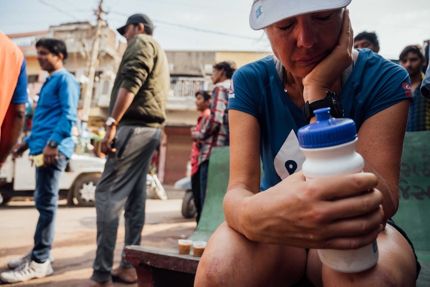 Mina Guli crouching, eyes closed holding her drink bottle while Indian pedestrians walk by in background