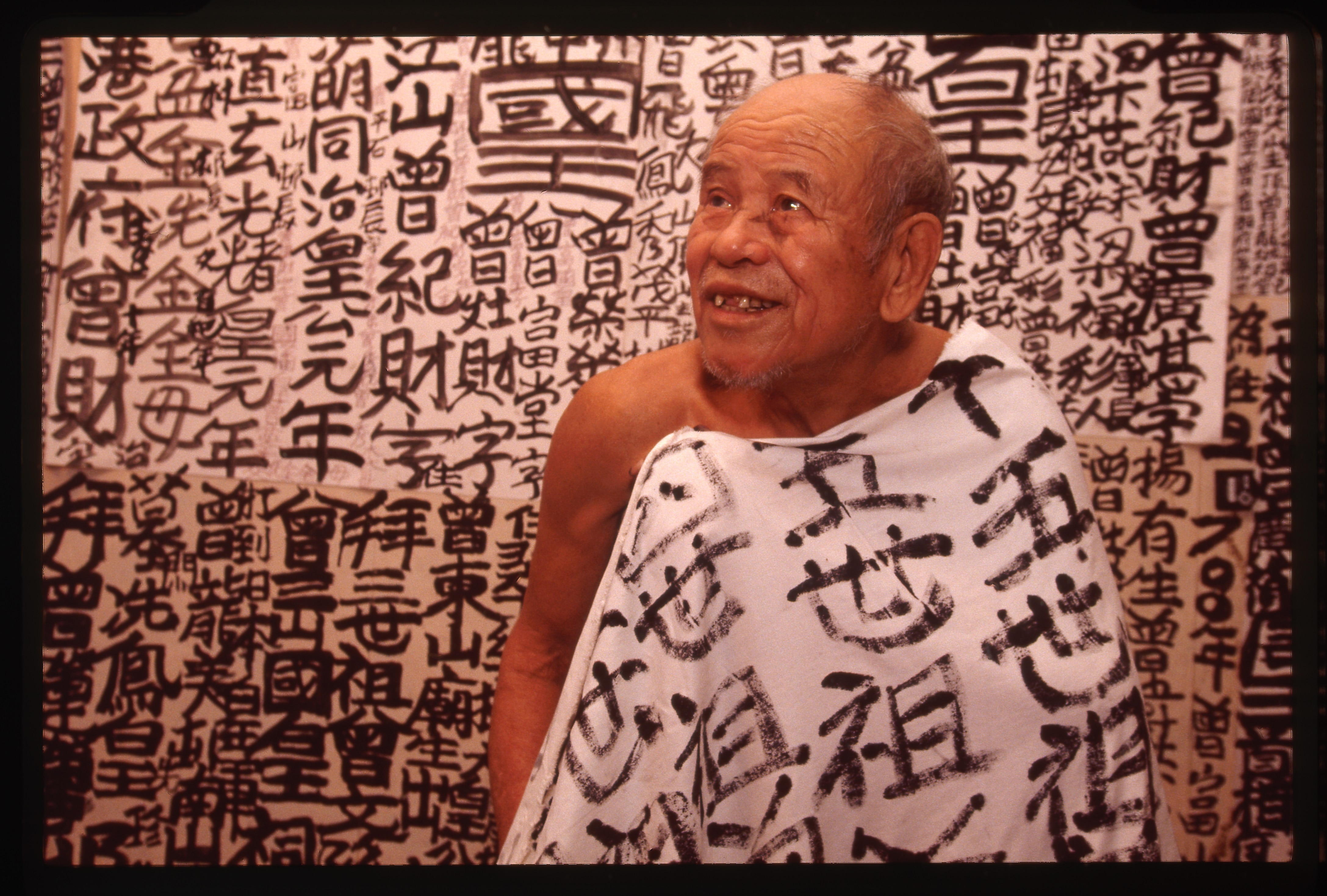 The King smiling draped in a sheet covered in his calligraphy in front of a wall of his calligraphy
