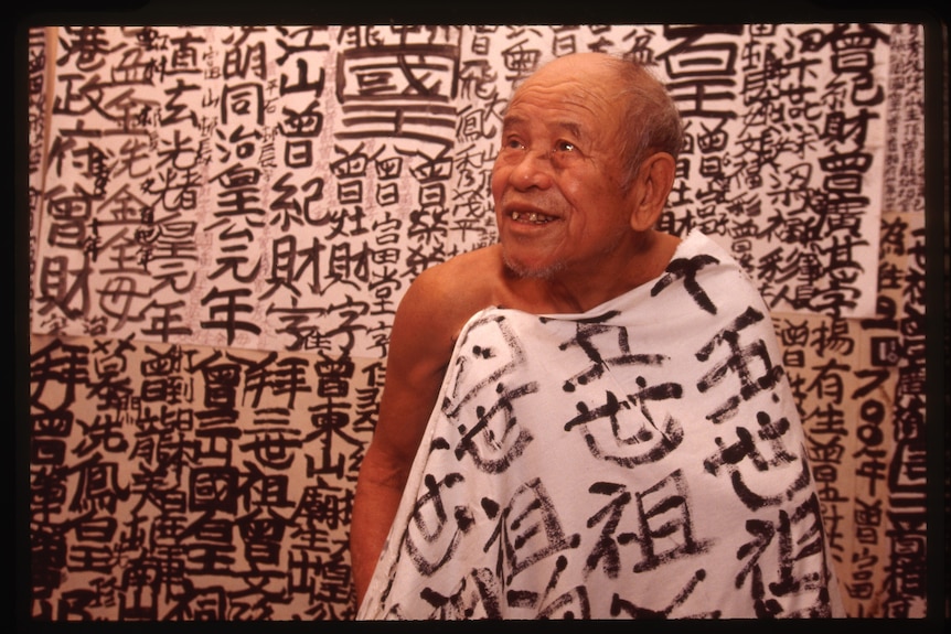 The King smiling draped in a sheet covered in his calligraphy in front of a wall of his calligraphy