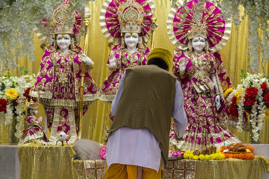 A man dressed in Indian clothes bends in front of tree heavily decorated Hindu deities. 