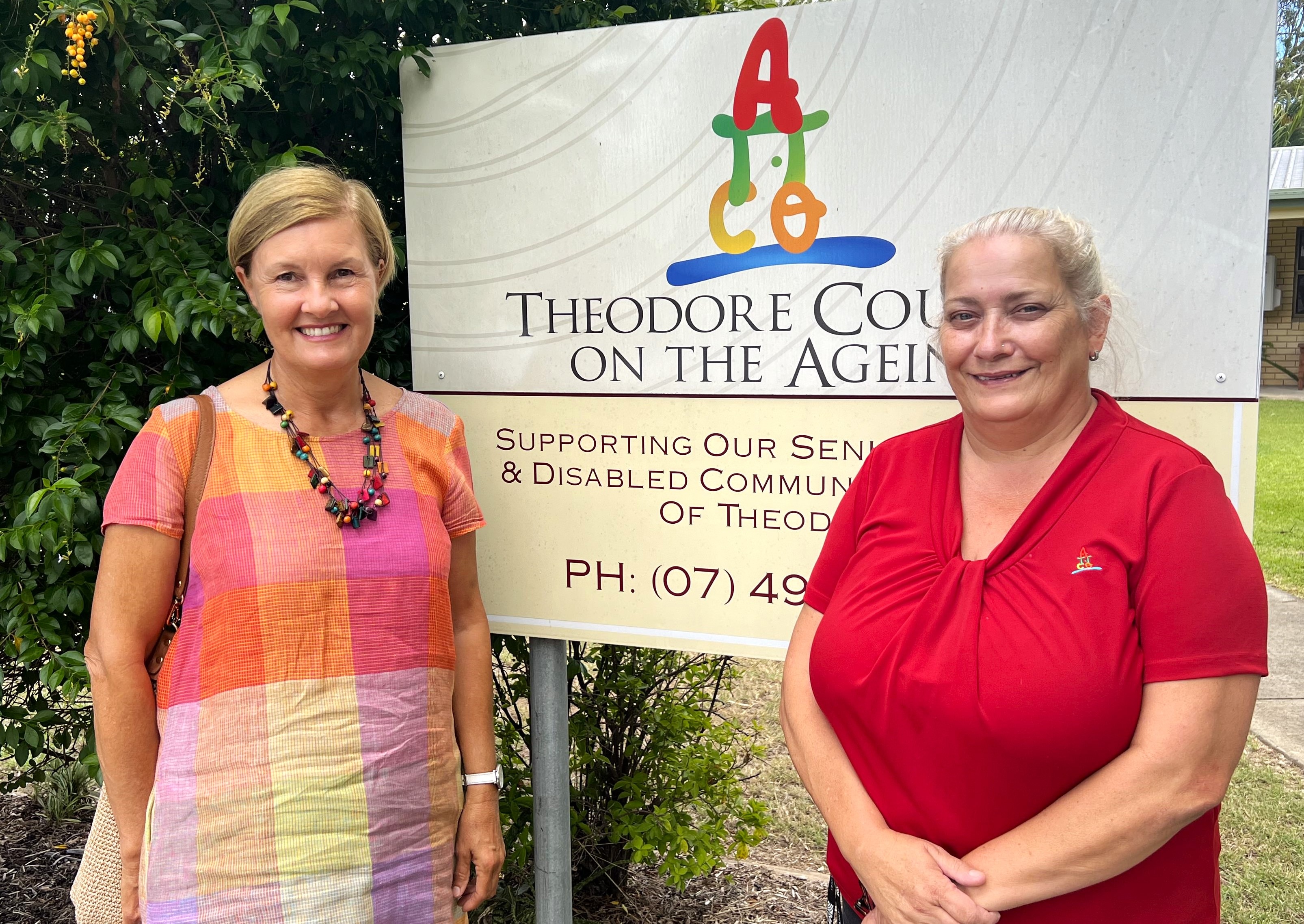 Two women dressed in bright colours smile standing next to Theodore Council on the Ageing sign