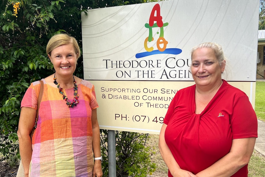 Two women dressed in bright colours smile standing next to Theodore Council on the Ageing sign