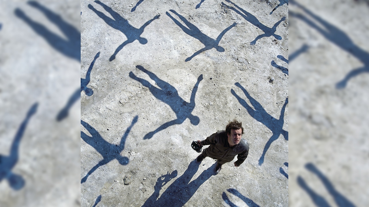 a man looks up to the sky, shadows of outstretched falling bodies surround him