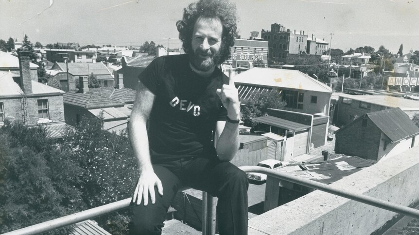 Black and white photo of Michael Gudinski in a Devo shirt, sitting on a rooftop and holding up his index finger