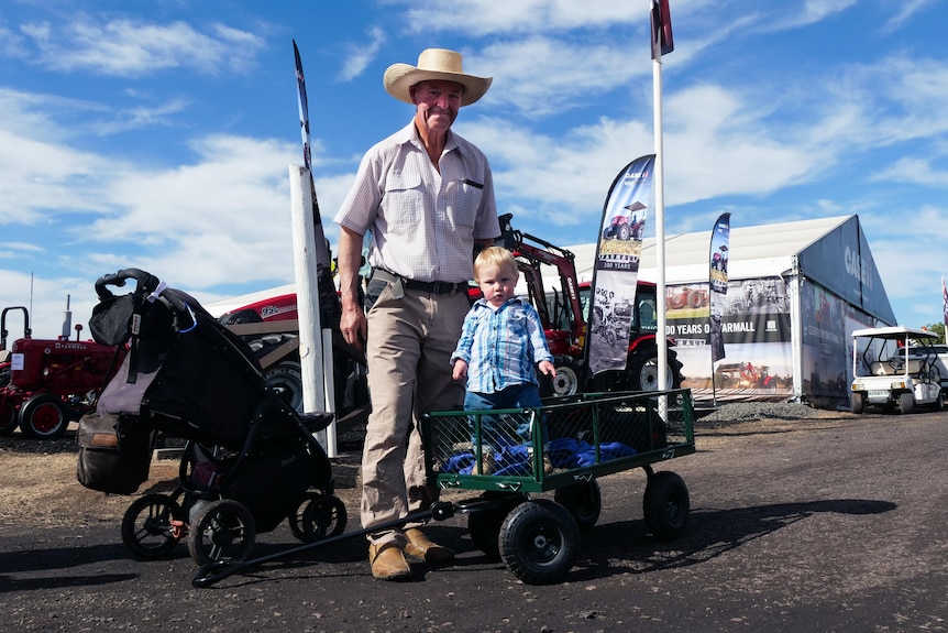 An older gentleman with an akubra stands next to his young grandson who is being towed in a small green trailer.