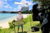 Andrew Greene standing under reflector boards in front of TV camera on beach in Guam.
