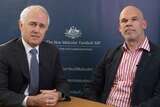 Malcolm Turnbull clasps his hands together on a desk. He sits next to Paul Shetler. Both men are wearing suits.