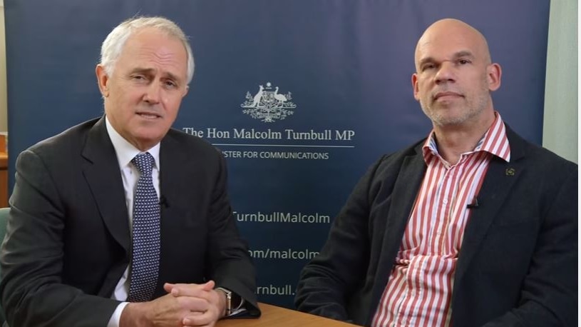 Malcolm Turnbull clasps his hands together on a desk. He sits next to Paul Shetler. Both men are wearing suits.