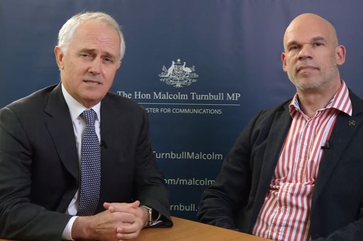 Malcolm Turnbull clasps his hands together on a desk. He sits next to Paul Shetler. Both men are wearing suits.