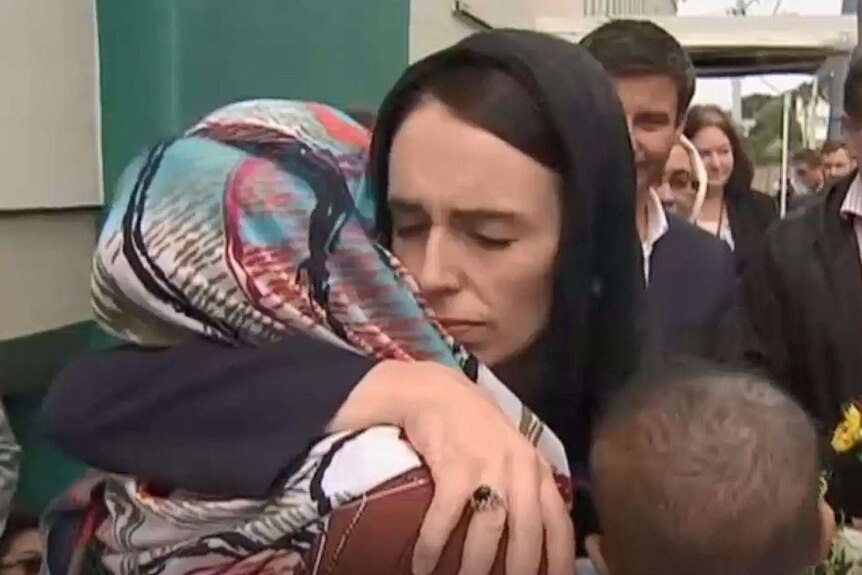 Jacinda Ardern has her arms around a woman in a hijab and is surrounded by a group of people.