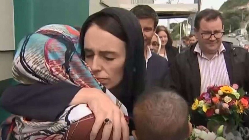 Jacinda Ardern has her arms around a woman in a hijab, and is surrounded by a group of people.