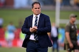 A serious looking rugby union coach clasps his hands together as he stands on the pitch after an international match.