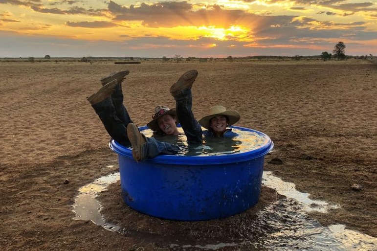 Two farm workers sit laughing, fully clothed, with their legs in the air, in a large tub of water on a dusty property.