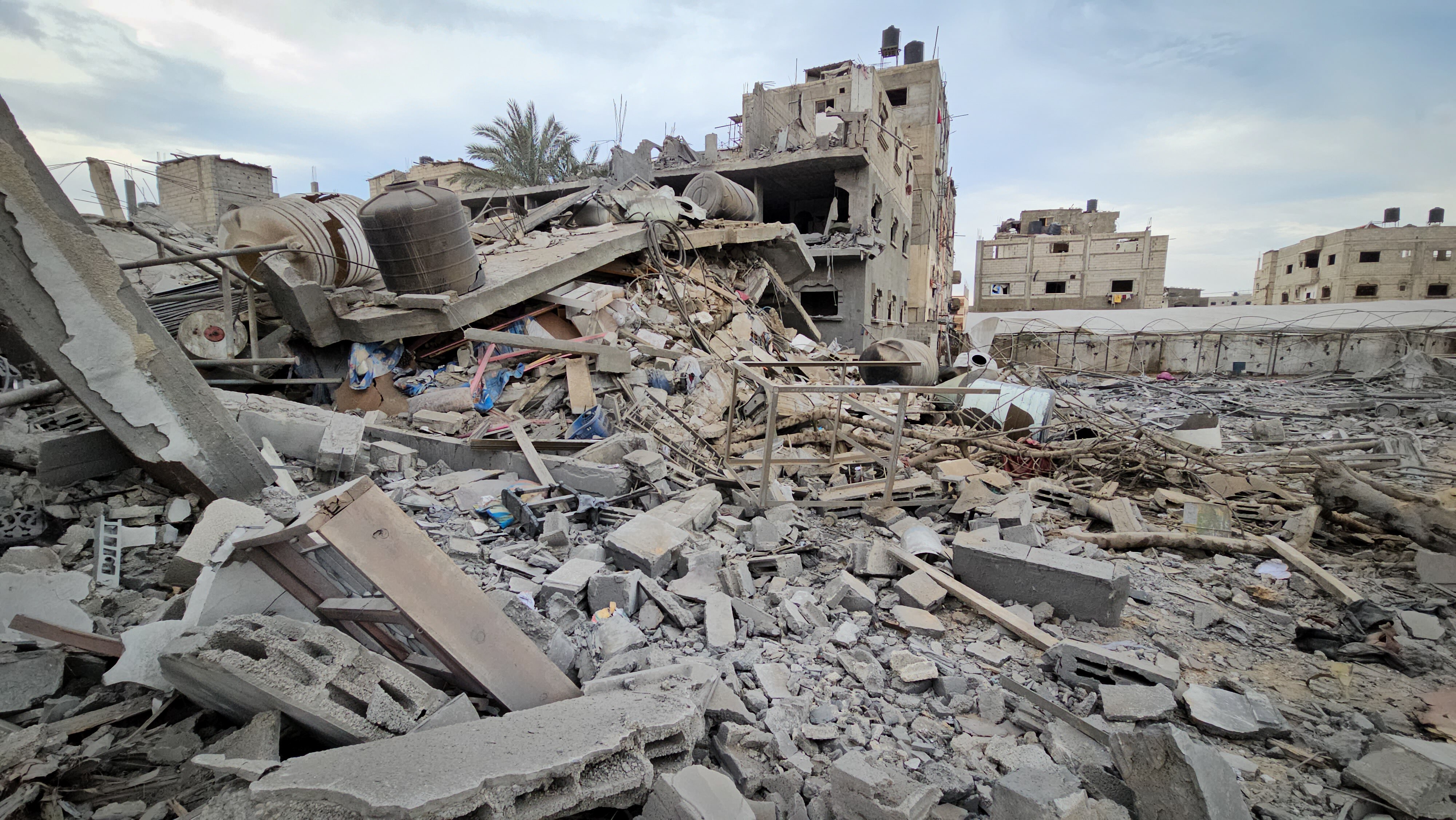 A mess of concrete and metal rubble on the ground, partially destroyed buildings behind