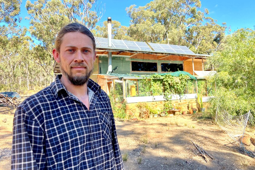 a photo of guy in flannel shirt standing in front of house with solar panels 