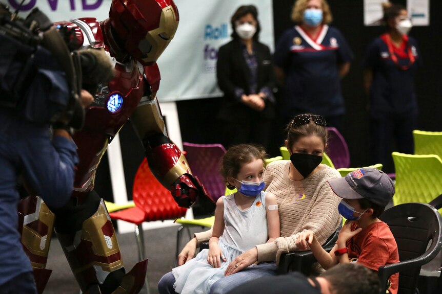 A Marvel superhero leans in towards children sitting down with their mum at a vaccination clinic.