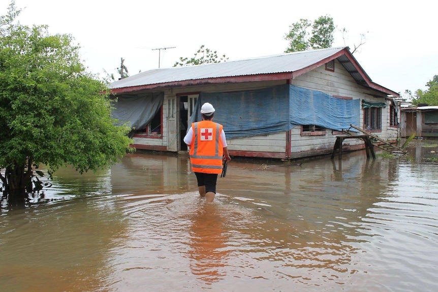 A Red Cross worker walks through knee-high floodwaters towards a house.