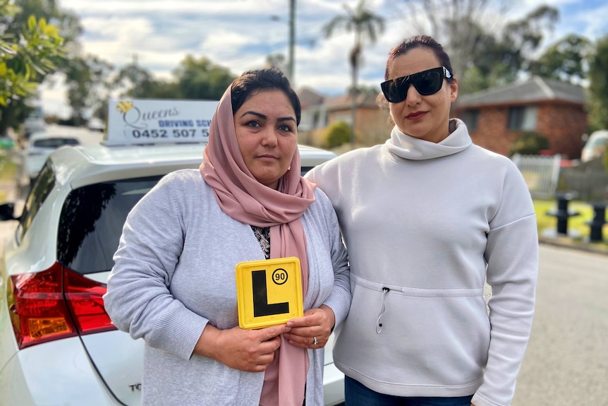 A woman holds an L-plate next to another woman in front of a car