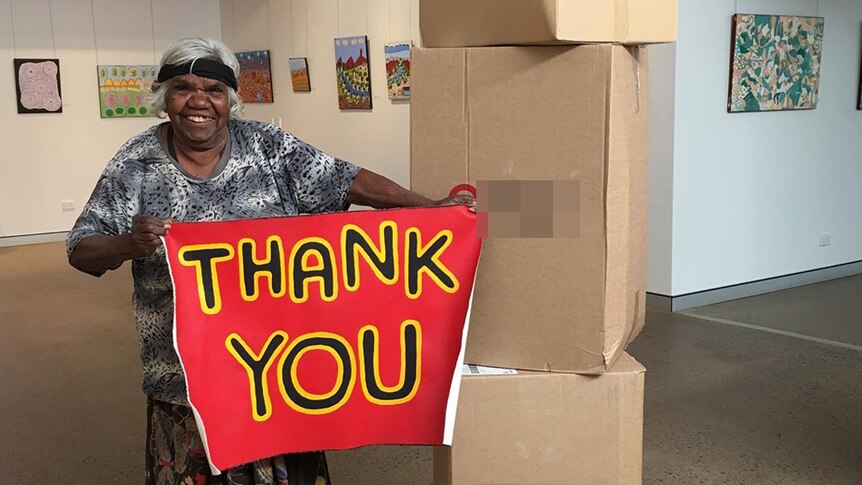 A grinning woman holds up a hand-painted sign that says 'thank you' next to a stack of boxes inside an art gallery.