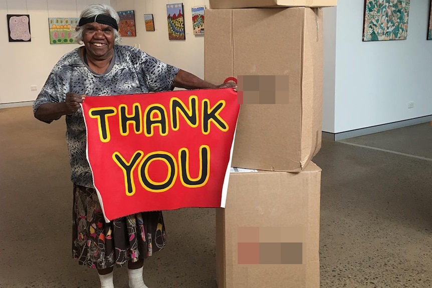 A grinning woman holds up a hand-painted sign that says 'thank you' next to a stack of boxes inside an art gallery.
