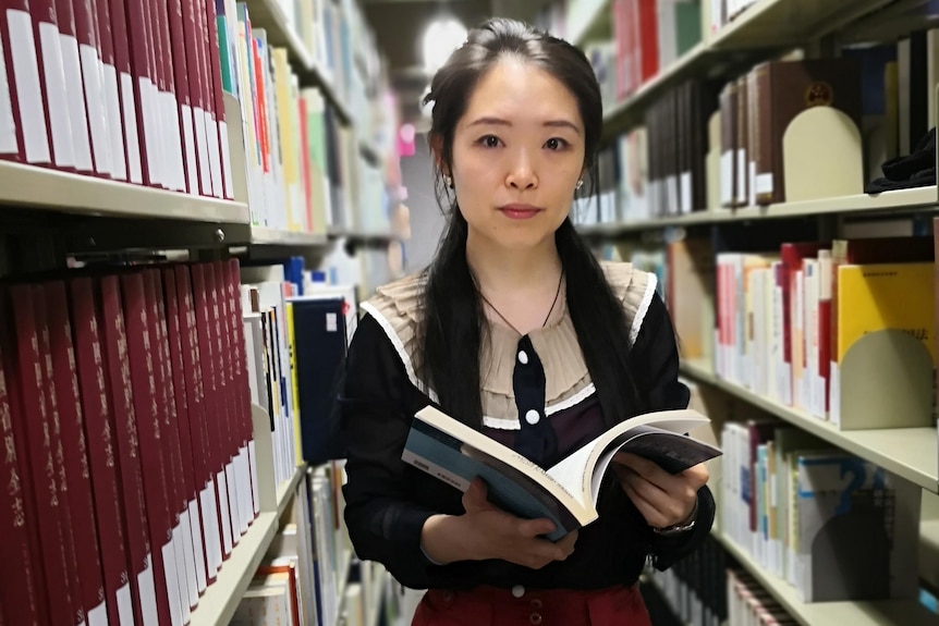 A woman holds a book open as she stands in an aisle of a library.