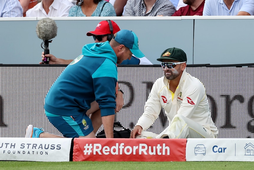 Nathan Lyon looks upset as he sits with a team doctor outside the boundary rope