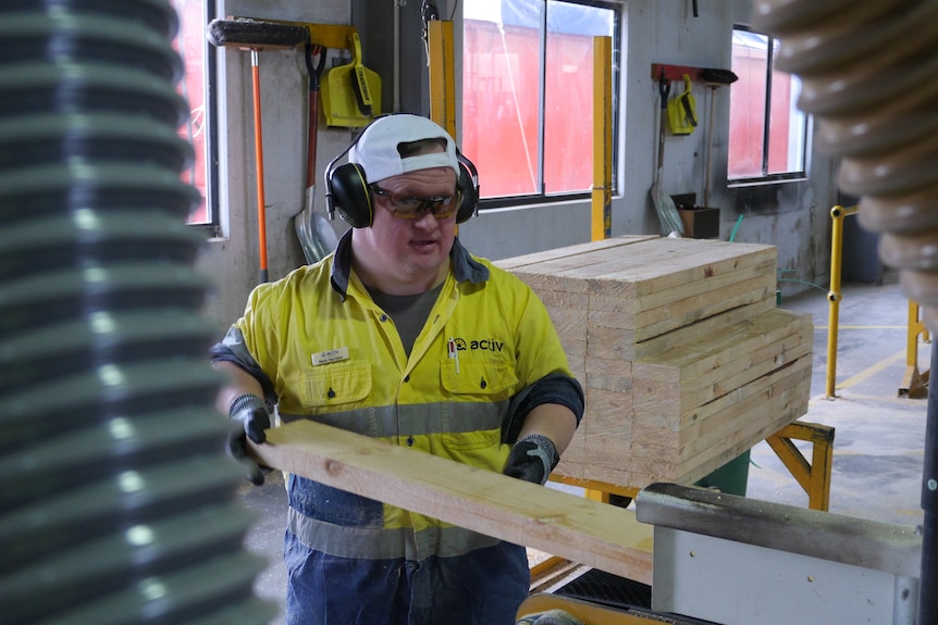 A man in high-vis wear, white cap and safety glasses saws a piece of wood