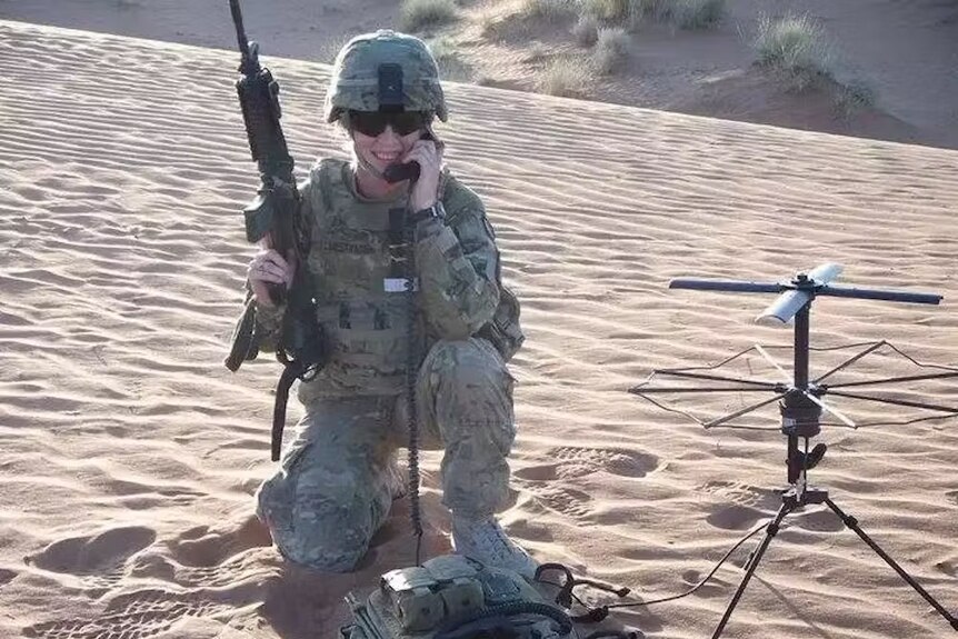 A woman in an army uniform and helmet poses for a photo on a beach, holding a gun and phone