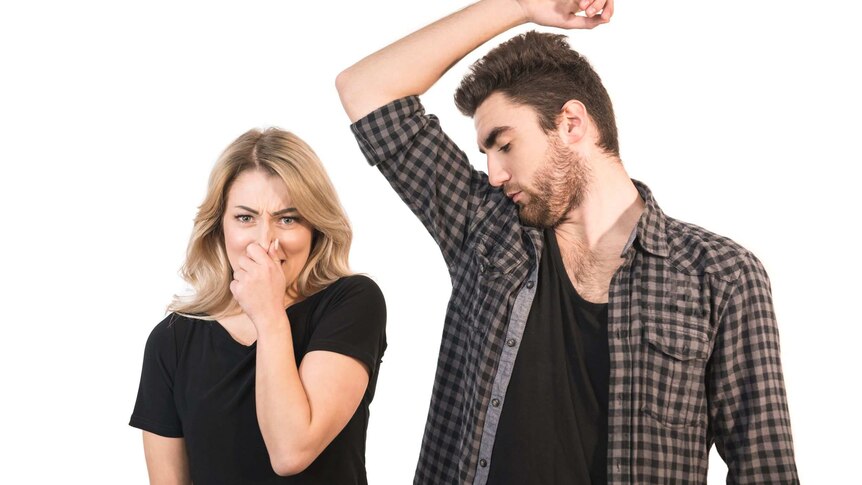 A woman with blonde hair holding her nose next to a bearded man wearing a checked shirt smelling his armpit