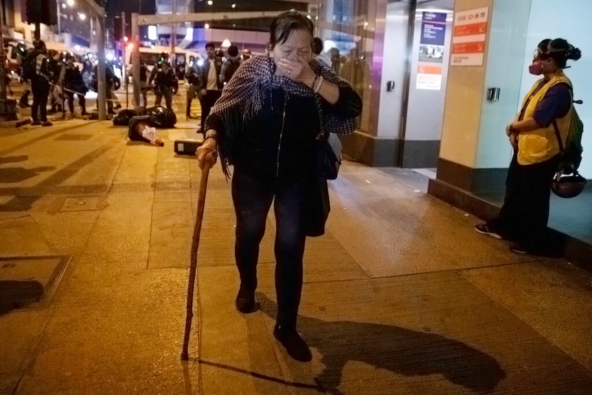 An elderly woman covers her mouth as she walks on a street with a walking stick with riot police gathering behind her.