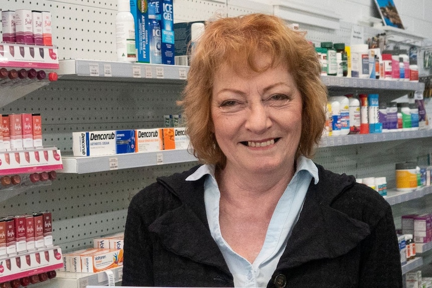 A woman with curly hair stands in a pharmacy, smiling.