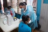 A nurse in full COVID PPE gives a vaccine shot to a seated woman wearing a face mask.