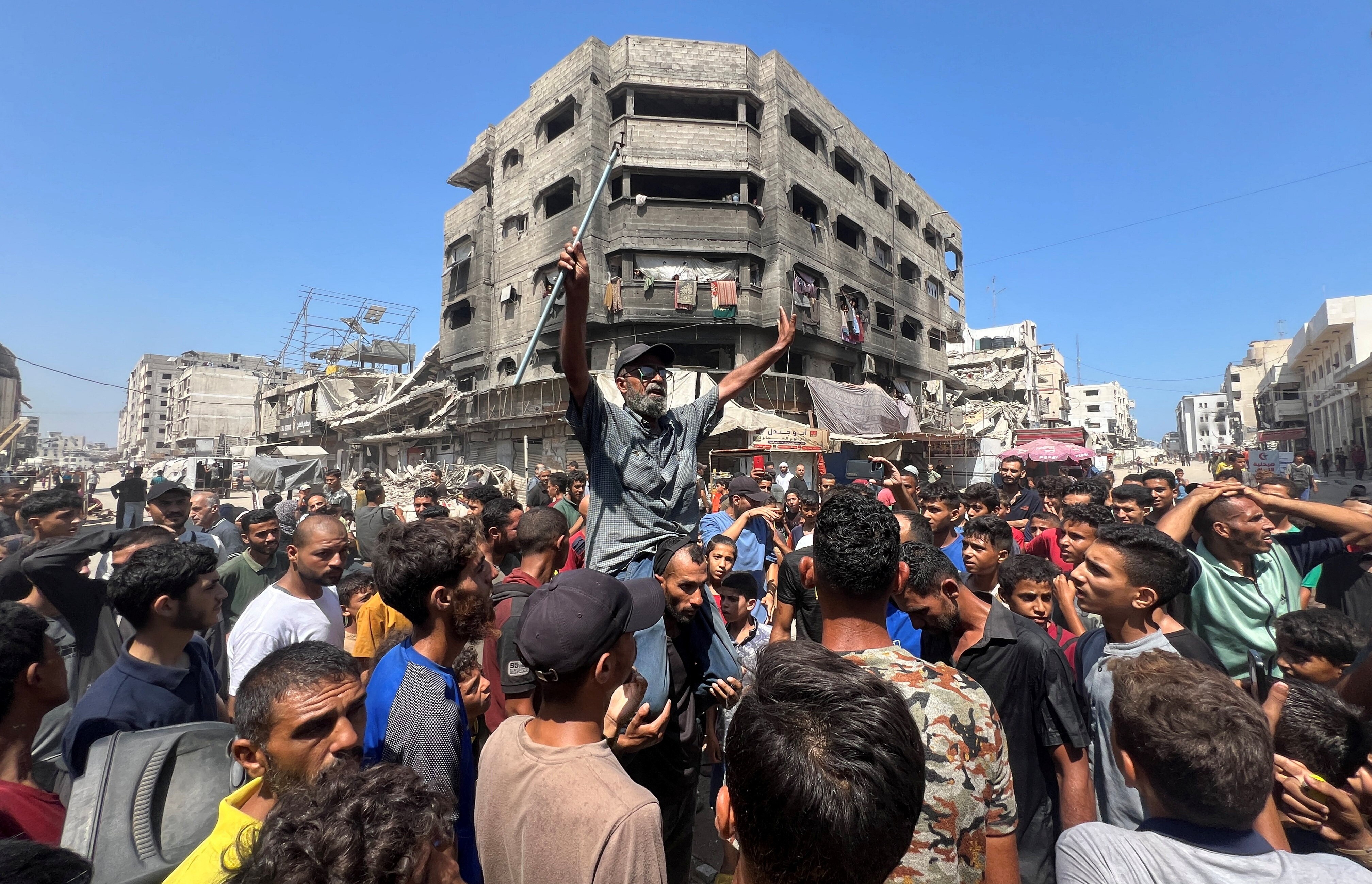 A man sits on the shoulders of another in the middle of a crowd with his arms out. A damaged building is in the background.